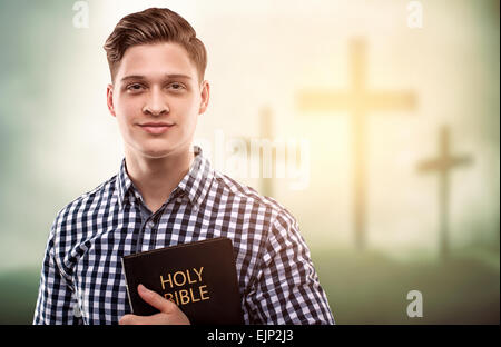 Schöner junger glücklicher Mann hält eine Bibel mit Baum kreuzt Hintergrund (Ostern-Konzept) Stockfoto