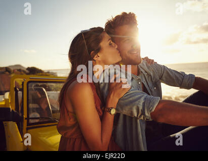 Romantische junges Paar sitzt auf der Motorhaube ihres Autos auf einem Roadtrip. Schöne junge Frau küssen ihren Boyfriend-look Stockfoto
