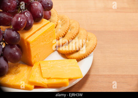 Cheddar-Käse, runde Kekse und roten Trauben sind auf einem kleinen weißen Teller mit einem Butcherblock Tischplatte unterhalb angeordnet. Stockfoto