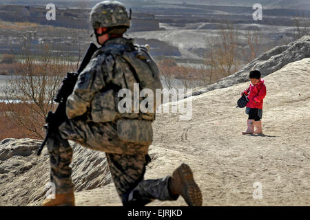 US Armee Sgt. Kevin Dixon hält eine Sicherheit auf dem Prüfstand, wie ein junges Mädchen auf die neue Winterjacke erhielt sie im Rahmen einer Bruderschaft Mission schaut, bekannt als ein Wroowali, unter der Leitung von afghanischen Regierungsbeamte und Polizisten in Bakorzai Dorf, Afghanistan, 22. Dezember 2010. Dixon ist der provinzielle Rekonstruktion Team Zabul Sicherheitstruppe zugeordnet. Staff Sgt Brian Ferguson Stockfoto