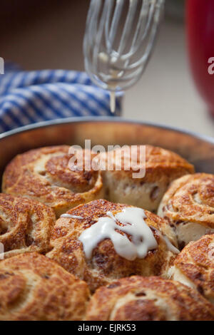 Zimtschnecken sitzen auf einem blau-weißen aufgegebenes Tuch, frisch aus dem Ofen. Eine Glasur Glasur tropft auf sie. Stockfoto