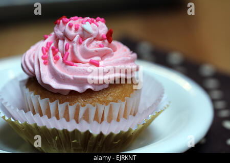 Ein Cupcake mit rosa Zuckerguss und Streusel rot, weiß und rosa sitzt auf einem weißen Teller über ein Braun eine weiße Serviette Stockfoto