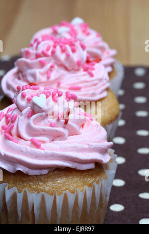 Drei Muffins auf einem braunen und weißen Tupfen Hintergrund mit rosa Zuckerguss und Streusel rosa, rot und weiß Stockfoto