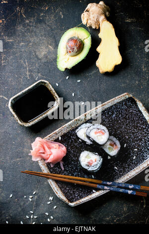 Sushi-Rollen mit Lachs, serviert auf schwarze Platte mit eingelegtem Ingwer, Avocado und Sojasauce und Stäbchen auf dunklem Hintergrund. T Stockfoto