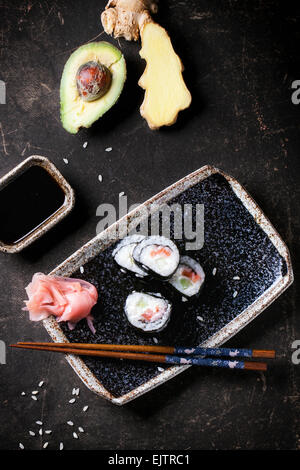 Sushi-Rollen mit Lachs, serviert auf schwarze Platte mit eingelegtem Ingwer, Avocado und Sojasauce und Stäbchen auf dunklem Hintergrund. T Stockfoto