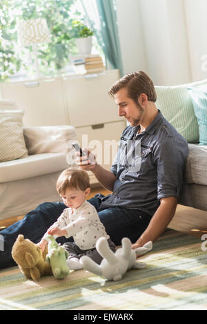 Vater sitzt auf Boden mit kleinen Sohn (2-3 Jahre) in Wohnzimmer und SMS-Nachricht Stockfoto