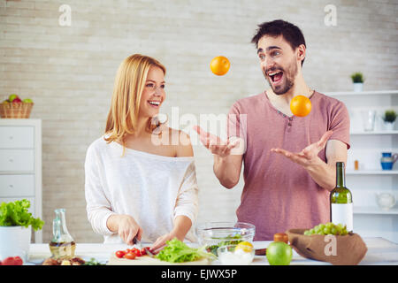 Glückliche junge Frau Kochen Salat in der Küche, gut aussehender Mann jonglieren Orangen in der Nähe von Stockfoto