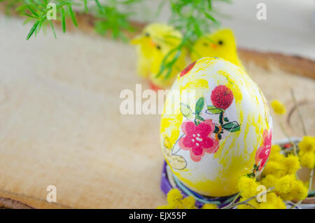 Handgemalte Decoupage Osterei auf einem Ständer mit gelben Blüten und Hühnern im Hintergrund auf einem Holztisch Stockfoto