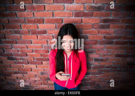 Glückliche Frau mit Smartphone über Ziegelmauer. Sportliche Jacke tragen Stockfoto