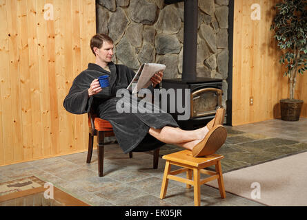 ein erwachsener Mann relaxen im Gewand und lese Zeitung mit Füßen Stockfoto