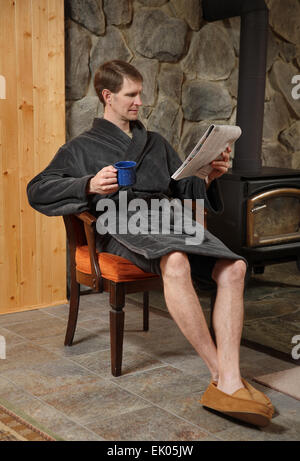 erwachsener Mann im Bademantel entspannen und lesen Zeitung Kamin mit Kaffeetasse Stockfoto