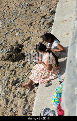 Aberystwyth, Wales, UK. 5. April 2015. Ostersonntag. Das Wetter. Zwei Frauen mit einem Smartphone auf der Meer-Betonwand neben dem Strand die Sonne genießen. Bildnachweis: Alan Hale/Alamy Live-Nachrichten Stockfoto