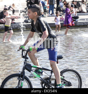 Bradford, West Yorkshire, Großbritannien, 6. April 2015. Erwachsene, Kleinkinder und Jugendliche genießen die Stadtpark mirror Pool und Brunnen im Centenary Square, Großbritanniens größte Stadtzentrum Wasserspiel. Asiatische junge auf Fahrrad im Pool, durchnäßt. Bildnachweis: Mick Flynn/Alamy Live-Nachrichten Stockfoto