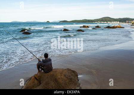 Mann sitzt auf einem Felsen an Palmas Strand Angeln. Governador Celso Ramos, Santa Catarina, Brasilien. Stockfoto