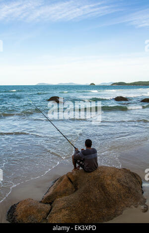 Mann sitzt auf einem Felsen an Palmas Strand Angeln. Governador Celso Ramos, Santa Catarina, Brasilien. Stockfoto