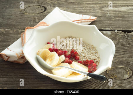 Haferflocken, Banane und gefrorene Erdbeeren in weißer Schale, Löffel auf rustikalen Tisch Stockfoto