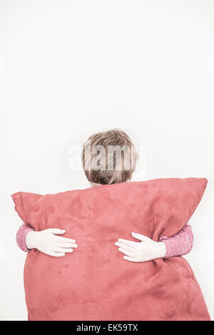 Trauriges Mädchen versteckt sich hinter einem großen roten Kissen. Sie steht vor einer weißen Wand mit viel Leerraum, die sein können Stockfoto