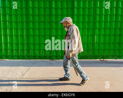 Eine männliche Hispanic Seniorin mit einem Bart und trägt eine Baseballkappe Typ, eine verschmutzte Jacke und blaue Jeans geht mit einem Stock werden Stockfoto