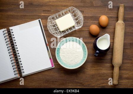 Zutaten für das Backen von Kuchen (Eiern, Mehl, Milch, Butter) auf Vintage Holztisch mit Rezeptbuch Stockfoto