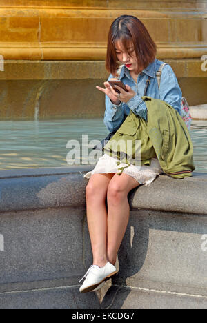 London, England, Vereinigtes Königreich. Japanische Frau sitzen von einem Brunnen am Trafalgar Square, SMS auf ihr Handy Stockfoto