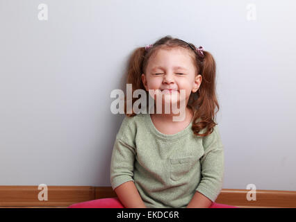 Fröhliches aufgeregt Mädchen Kniff ihre Augen und warten etwas gutes auf dem Boden sitzend Stockfoto