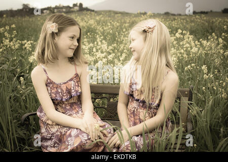 Zwei Schwestern Lachen gemeinsam beim Sitzen auf einer Bank in einem Feld von gelben Blüten. Stockfoto