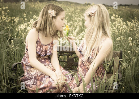 Zwei Schwestern sitzen zusammen auf einer Bank in einem Feld von gelben Blüten. Stockfoto