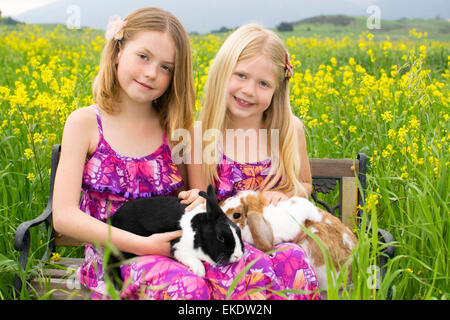 Zwei schöne Schwestern halten Hasen sitzend auf einer Bank in einem Feld von gelben Blüten. Stockfoto