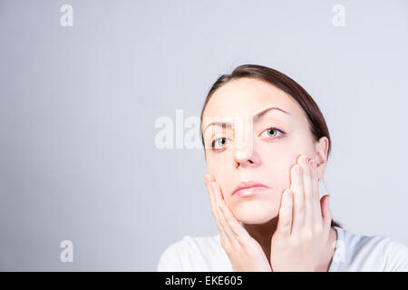 Nahaufnahme ernste junge Frau berühren ihr Gesicht mit beiden Händen während in die Kamera schaut. Im Studio mit leichten grauen Ba erfasst Stockfoto