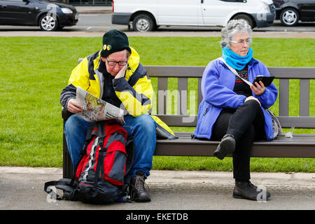 Mann und Frau sitzen auf öffentlichen Bänken im Stadtzentrum von Glasgow, man liest eine Zeitung, die andere lesen ihr Handy, Glasgow Stockfoto