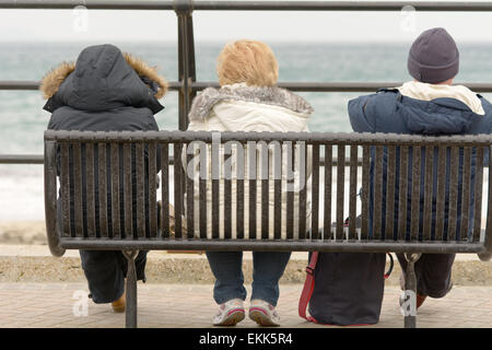 Drei Touristen ihren Urlaub saß auf der Bank, Blick auf das Meer an kalten bewölkten Frühlingstag in Looe, Cornwall, England Stockfoto