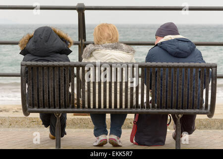 Drei Touristen ihren Urlaub saß auf der Bank, Blick auf das Meer an kalten bewölkten Frühlingstag in Looe, Cornwall, England Stockfoto