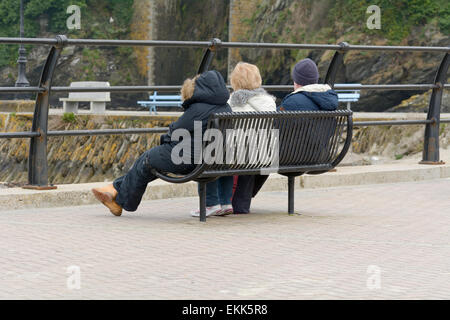 Drei Touristen ihren Urlaub saß auf der Bank, Blick auf das Meer an kalten bewölkten Frühlingstag in Looe, Cornwall, England Stockfoto