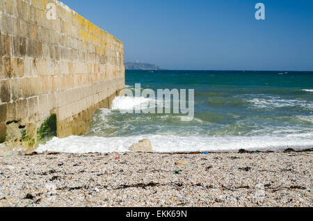 Seascape Bild mit Kiesstrand, Hafen Wand Mole, Leuchtturm in der Ferne und blauer Himmel Stockfoto