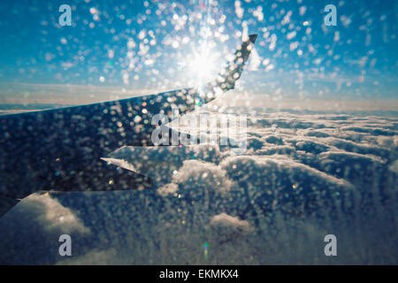 Blick auf Flugzeugflügel und Wolken von Innenkabine an sonnigen Tag mit Eis auf dem Fenster. Stockfoto