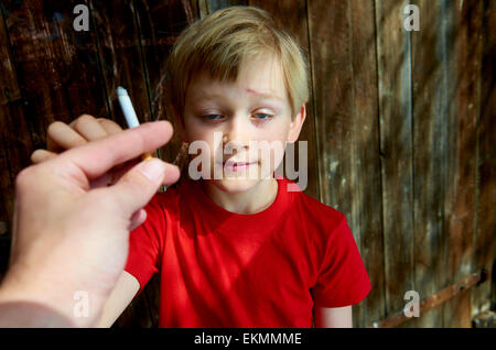 Porträt des Kindes Blondschopf qualmender Zigarette mit dunklem Holz Hintergrund hinter ihm. Erwachsener Mann Hand junge eine Zigarette anzubieten Stockfoto