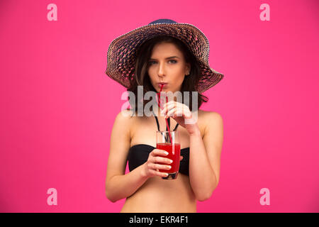 Junge Frau im Bikini und Hut trinken Saft über rosa Hintergrund Stockfoto