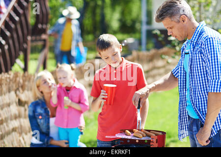 Junger Mann Braten Würstchen auf dem Grill im Freien mit seinem Sohn in der Nähe von Stockfoto