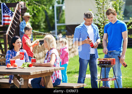 Zwei Männer auf Würstchen braten Grillen im Freien mit jungen Frauen reden vom Tisch in der Nähe von Stockfoto