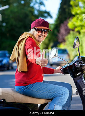 Trendige Großmutter in einer Schirmmütze Schal und Jeans ein Motorroller entlang der Straße und Lachen mit Genuss beim geben einer t Stockfoto