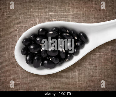 Schwarze Bohnen in weißem Porzellanlöffel / Highres Foto von Korn in weißem Porzellan Löffel auf Jute Sackleinen Hintergrund Stockfoto