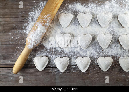 Herzförmige Knödel, Mehl und Nudelholz auf hölzernen Hintergrund. Ravioli kochen. Ansicht von oben. Stockfoto