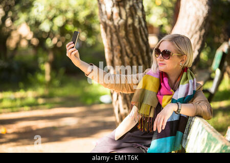 Kaukasische Mädchen Lächeln für ein Selbstportrait (Selfie) auf ihrem Smartphone, sitzen auf der Bank im Park. Stockfoto