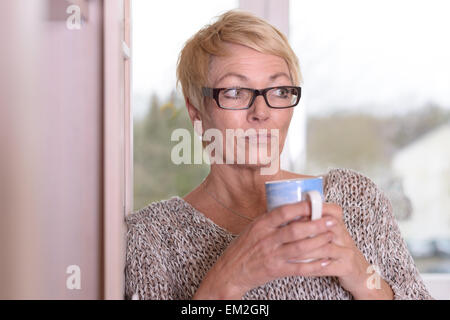 Nahaufnahme der schweren Mittelalter Frau tragen Brillen, mit kurzen blonden Haaren, hält eine Tasse während lehnen an der Wand und auf der Suche Stockfoto