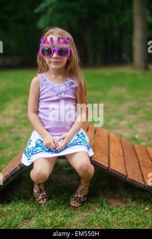 Niedliche kleine Mädchen in lila Happy Birthday Brille auf hölzernen Stuhl im freien Stockfoto