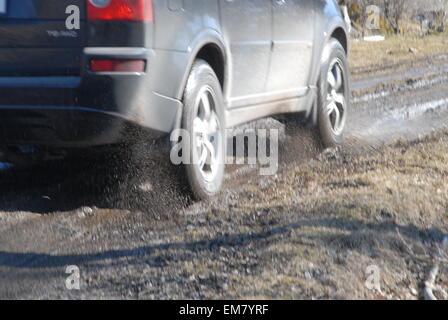 Fahrzeugräder spritzen Wasser beim Fahren durch Pfützen auf einer überfluteten Schlammstraße. Stockfoto