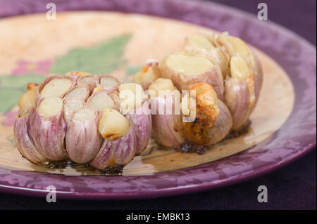 Zwei gebratene lila Streifen Knoblauch Zwiebeln, zeigen die einzelnen Zehen auf einem lila und tan Platte dekoriert Stockfoto