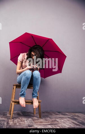 Frau sitzt auf dem Stuhl mit rosa Regenschirm und mittels Smartphone im Studio. In Jeans und t-Shirt tragen. Barfuß Stockfoto