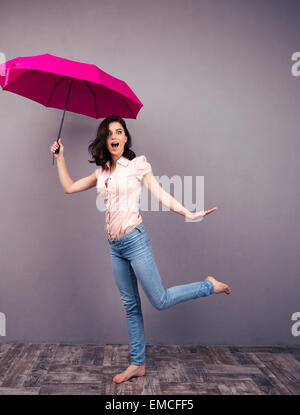 In voller Länge Portrait einer überrascht Frau posiert mit rosa Regenschirm im Studio. In Jeans und t-Shirt tragen. Barfuß. Mit Blick auf Stockfoto