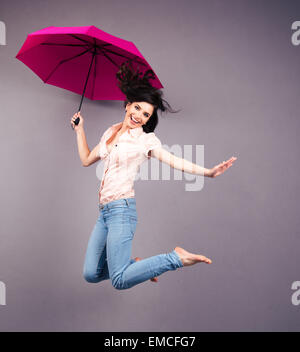 Glückliche junge Frau mit Regenschirm über grauen Hintergrund springen. Blick in die Kamera Stockfoto